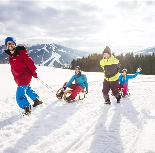 Eine Gruppe von Freunden zieht einen Schlitten im Schnee. Die Sonne scheint und die Berge sind im Hintergrund zu sehen.