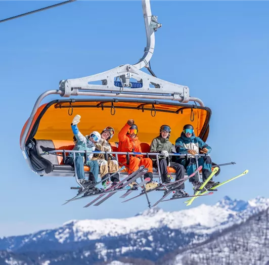 Eine Gruppe von Skifahrern sitzt fröhlich in einem Sessellift. Im Hintergrund sind schneebedeckte Berge und ein klarer blauer Himmel zu sehen.