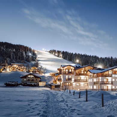 Das Hotel Lisa in Abendbeleuchtung direkt an der Gondelbahn in Flachau. Im Hintergrund sieht man eine beleuchtete Skipiste und einen klaren Nachthimmel.
