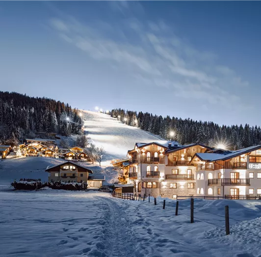 The Hotel Lisa in evening lighting right by the gondola lift in Flachau. In the background, you can see a lit ski slope and a clear night sky.