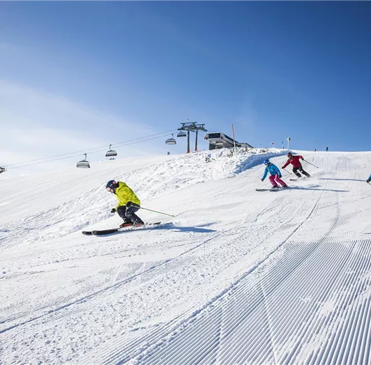 Skifahrer fahren über eine gut präparierte Piste im Schnee. Der Himmel ist klar und blau, und im Hintergrund sind Skilifte zu sehen.