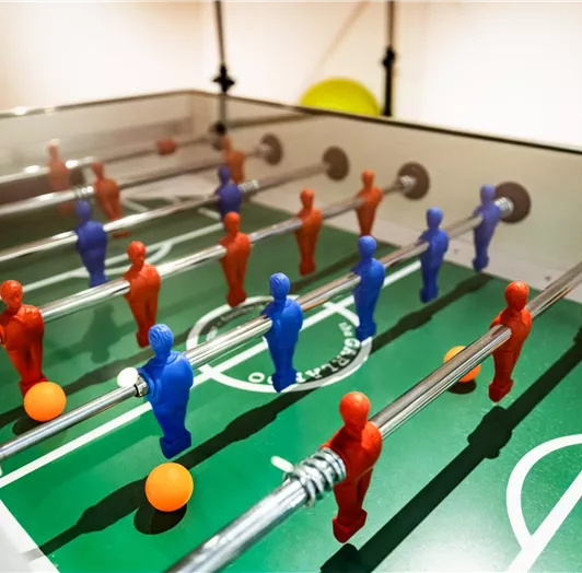 Ein Tischfußballspiel mit blauen und roten Figuren auf einem grünen Spielfeld. Es gibt auch orangefarbene Bälle, die im Spiel verwendet werden.