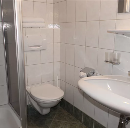 Ein modernes Badezimmer mit weißen Fliesen und einer Dusche. Es gibt ein Waschbecken, eine Toilettenschüssel und ein Regal mit Handtüchern.