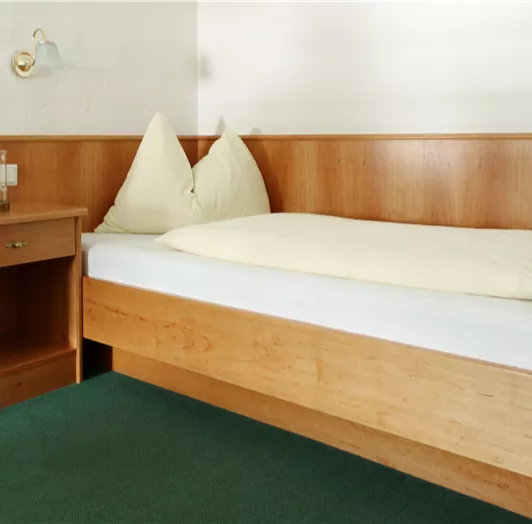 Ein einfaches Schlafzimmer mit einem Einzelbett und zwei Kissen. Neben dem Bett steht ein Nachttisch mit einer Lampe und einem Glas.