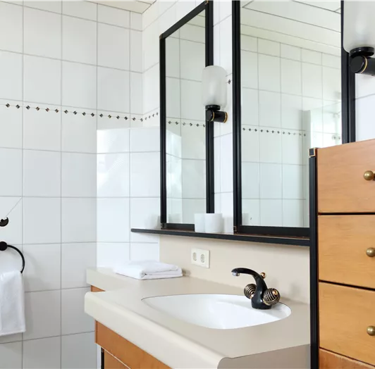 Ein modernes Badezimmer mit weißen Fliesen und einem Waschbecken. Es gibt einen Spiegel, einen Handtuchhalter und eine Holzkommode mit Schubladen.