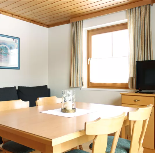 Ein helles Wohnzimmer mit einem Esstisch und Stühlen aus Holz. Im Hintergrund sind eine Couch, ein Fernseher und ein Fenster mit Vorhängen zu sehen.