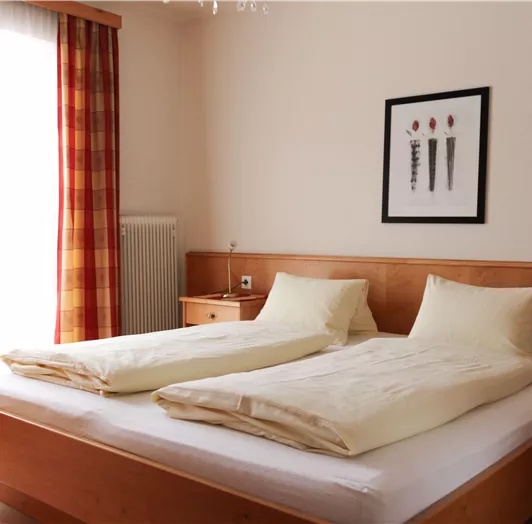 Ein modernes Schlafzimmer mit einem Doppelbett und hellen Kissen. Große Fenster lassen viel Licht herein und es gibt ein dekoratives Bild an der Wand.