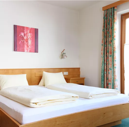Ein helles Schlafzimmer mit einem Doppelbett und einer Holzkommode. Große Fenster lassen viel Tageslicht herein und die Wände sind mit einem bunten Bild und Vorhängen dekoriert.