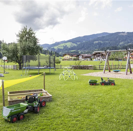 Ein Spielplatz mit einer Wiese, auf der Spielzeuge wie Traktoren stehen. Im Hintergrund sind Schaukeln und Berge zu sehen.