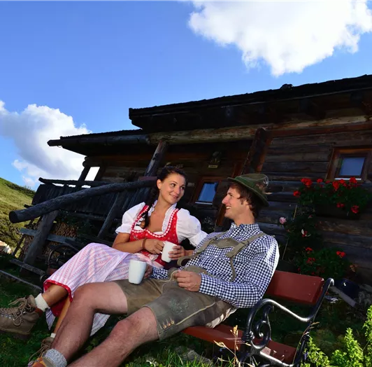 Ein Paar in traditioneller Tracht sitzt entspannt vor einer Holzhütte.