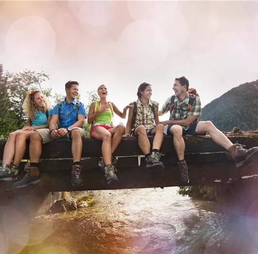 Eine Gruppe von Freunden sitzt fröhlich auf einer Holzbrücke über einem Fluss.