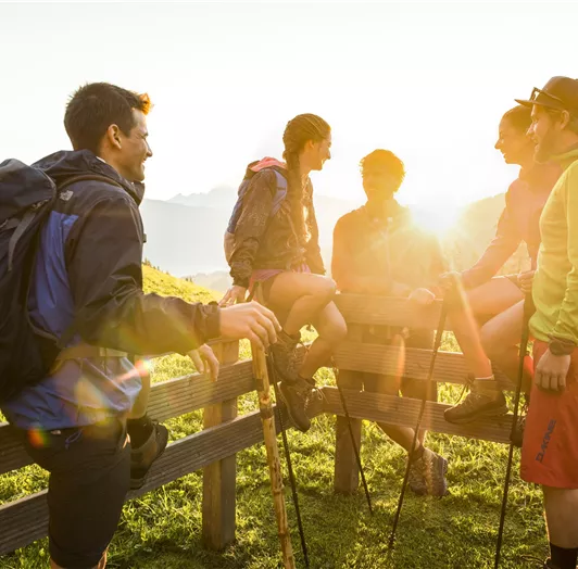Eine Gruppe von fünf Wanderern unterhält sich fröhlich an einem Holzzaun in der Natur.