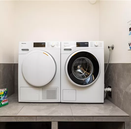 Eine moderne Waschmaschine und ein Wäschetrockner stehen nebeneinander in einem hellen Raum. Daneben liegt eine Packung Waschmittel auf einem grauen Tisch.