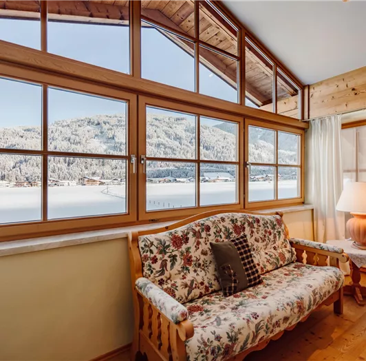 Studio on the upper floor: A cozy living room with a floral sofa and large windows. Outside, you can see a snow-covered landscape with mountains from Flachau.