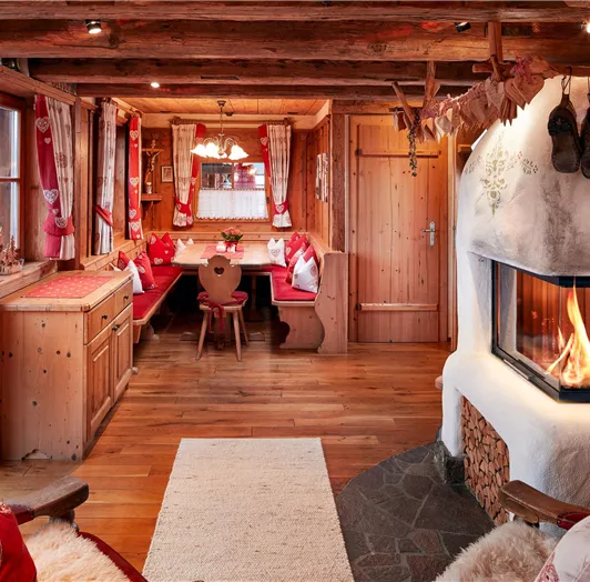 Uriges Wohnzimmer mit Kamin und traditioneller Eckbank. Behagliches Wohnzimmer im traditionellen Alpenstil mit modernem Komfort. Der gemütliche Kachelofen sorgt für wohlige Wärme und lädt zum Entspannen ein. Die große, umlaufende Eckbank mit weichen Polstern und herzverzierten Kissen bietet einen perfekten Platz für gemeinsame Mahlzeiten oder gesellige Stunden mit Familie und Freunden. Rustikale Holzbalken, natürliche Materialien und liebevolle Details wie bestickte Vorhänge, Holzschnitzereien u